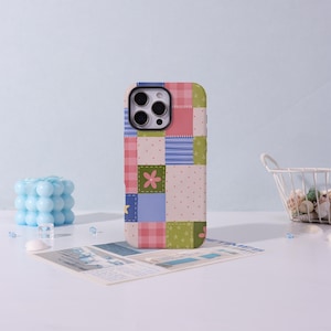 Könnte beinhalten: Eine Smartphone-Hülle mit einem bunten Patchwork-Design aus verschiedenen Quadraten in Rosa-, Blau-, Grün- und Cremetönen. Das Muster enthält kleine Blumenmotive, Punkte, Karos und Sternformen, die an eine Steppdecke erinnern.