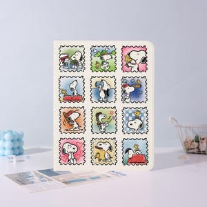 Könnte beinhalten: Ein rechteckiges Notizbuch mit einem Raster aus zwölf briefmarkenartigen Illustrationen von Snoopy und Woodstock. Jede einzelne Briefmarke zeigt die Figuren bei verschiedenen Aktivitäten wie Wandern, Lesen und Entspannen in sanften Farben.