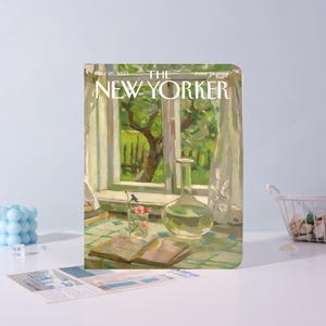 May include: A notebook with a cover featuring a painting of a window scene with a view of a green yard. The cover reads "THE NEW YORKER" with the date "May 26, 1923" and the price "15 cents". A glass bottle and a small vase with flowers are on a table.