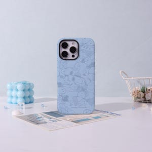 May include: A light blue phone case featuring a repeating pattern of cartoon characters. The case has a matte finish and is designed to fit a smartphone. The background includes a blue candle, a small basket, and a newspaper.