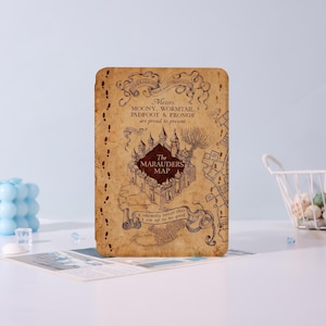 May include: A rectangular, tan-colored item featuring a detailed map design with the text "The Marauders Map." The map includes illustrations of a castle, footprints, and various text elements. The item has a vintage aesthetic.