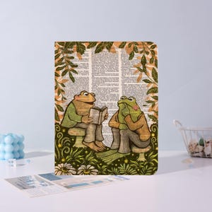 May include: A rectangular notebook cover featuring a printed illustration of two anthropomorphic frogs sitting on mushrooms in a forest. One frog reads a book while the other rests its chin on its hand. The background consists of dictionary text.
