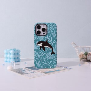 May include: A phone case featuring a black and white orca whale design. The case has a teal background with a pattern of white coral and seaweed. The orca is swimming in the center of the case. The phone case is a protective accessory.