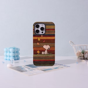 May include: A mobile phone case with a striped design in autumnal colours of brown, red, and orange. The case features a cartoon illustration of Snoopy and Woodstock. The case has a glossy finish.