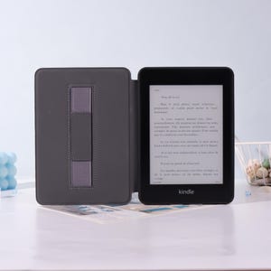 Puede incluir: Un lector de libros electr&oacute;nicos Kindle negro con una funda gris, abierto para mostrar texto. La funda tiene una correa gris para sujetar el dispositivo. El Kindle est&aacute; sobre una superficie blanca.