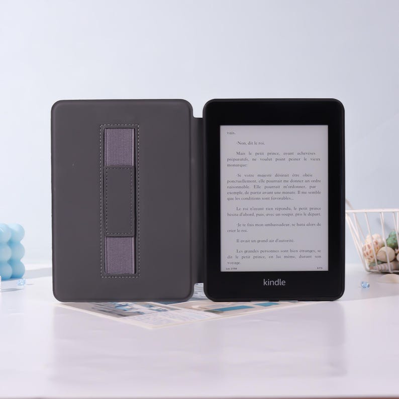 Puede incluir: Un lector de libros electr&oacute;nicos Kindle negro con una funda gris, abierto para mostrar texto. La funda tiene una correa gris para sujetar el dispositivo. El Kindle est&aacute; sobre una superficie blanca.