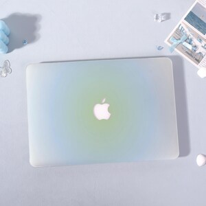 May include: A laptop computer with a gradient blue and green cover, featuring the Apple logo in the center. The cover has a smooth, matte finish. The laptop is on a light blue surface.