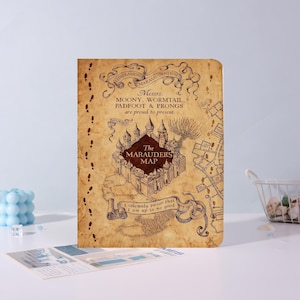 May include: A rectangular notebook with a tan, parchment-style cover featuring a dark brown illustration of a castle and the text "The Marauders Map," "Messrs. Moony, Wormtail, Padfoot & Prongs are proud to present," and "I solemnly swear that I am up to no good."