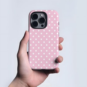 May include: A pink phone case featuring a white polka dot design. The case is held in a hand and has a camera aperture. The phone case is designed to fit a smartphone.