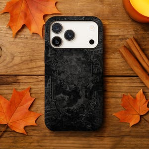 May include: Black phone case featuring a detailed, dark artwork design. The case has a camera cutout and a sleek, modern look. The background includes autumn leaves and a wooden surface.