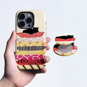 May include: A phone case and pop socket featuring a stack of colorful pillows with a black cat silhouette on top. The case is held in a hand, showcasing the design's vibrant red, yellow, and pink hues.