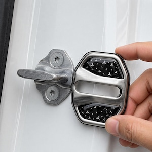 May include: A silver-colored car door lock cover with a black patterned insert featuring white star-like shapes. The cover is being held near the door latch, which is silver-colored. The background is a white car door.