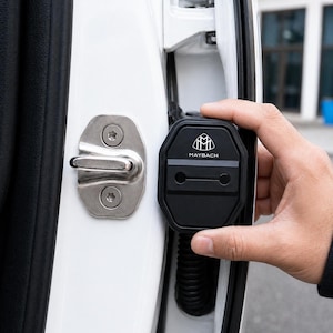 May include: Black car door lock cover with the Maybach logo. The cover is being held next to the door latch. The door is white.