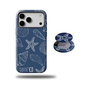 May include: A navy blue phone case and matching pop socket featuring a white seashell and starfish pattern. The phone case has a glossy finish and a camera cutout. The pop socket is round and has a matching design.