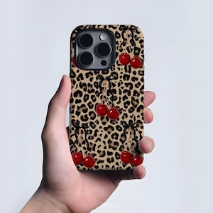 Op de afbeelding: Een telefoonhoesje met een luipaardprint en rode kersen vastgebonden met zwarte strikken. De hoes heeft een glanzende afwerking en een camera-opening. De achtergrond is lichtbeige.