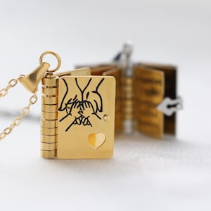 May include: A gold-coloured locket necklace shaped like a book. The front cover has a black line drawing of hands and a heart-shaped cutout. The locket is open, revealing pages inside.