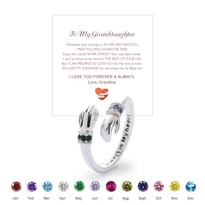 May include: Silver ring with two hand designs, one with green gemstones and the other with purple gemstones. The ring has the words "Always in My Heart" engraved on the band. A card with text "To My Granddaughter" is in the background.