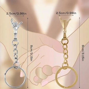 May include: Two keychains, each 8 inches (20 cm) long, featuring a hand gesture design. One keychain is silver-colored, the other gold-colored. Both have a chain and a round key ring. The hand gesture is 0.98 inches (2.5 cm) wide.