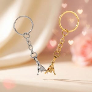 May include: Two keychains, one silver and one gold, each featuring a hand gesture charm. The charms are connected to a chain and a keyring. The background is a soft, blurred gradient with heart-shaped bokeh.