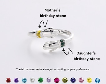 May include: A silver-colored ring with two hand-shaped ends, each adorned with a gemstone. One stone is yellow, the other green. Text on the ring reads "You". The image includes text indicating the ring's customizable birthstones.