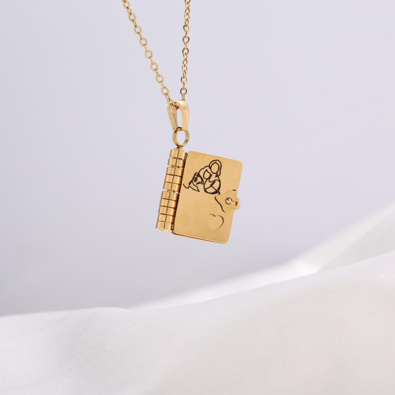 May include: A gold-colored pendant necklace featuring a small, book-shaped locket. The locket has a heart and a design on the front. The necklace chain and pendant are made of a shiny, gold-toned metal.
