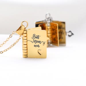 May include: A gold-coloured pendant necklace with a small, book-shaped locket. The locket is engraved with the words "Best Mom ever" and a heart. The necklace has a delicate chain and a clasp.