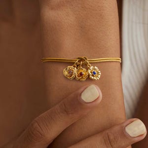 May include: A gold chain bracelet with three flower charms. The charms are made of gold and have red, blue, and pink gemstones in the center.