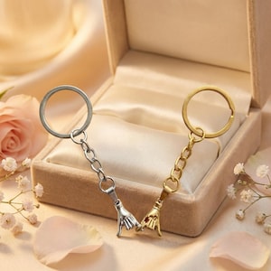 May include: Two keychains, one silver and one gold, each featuring a hand gesture charm. The charms are connected by a chain to a keyring. The keychains are presented in a small, open gift box on a satin surface.