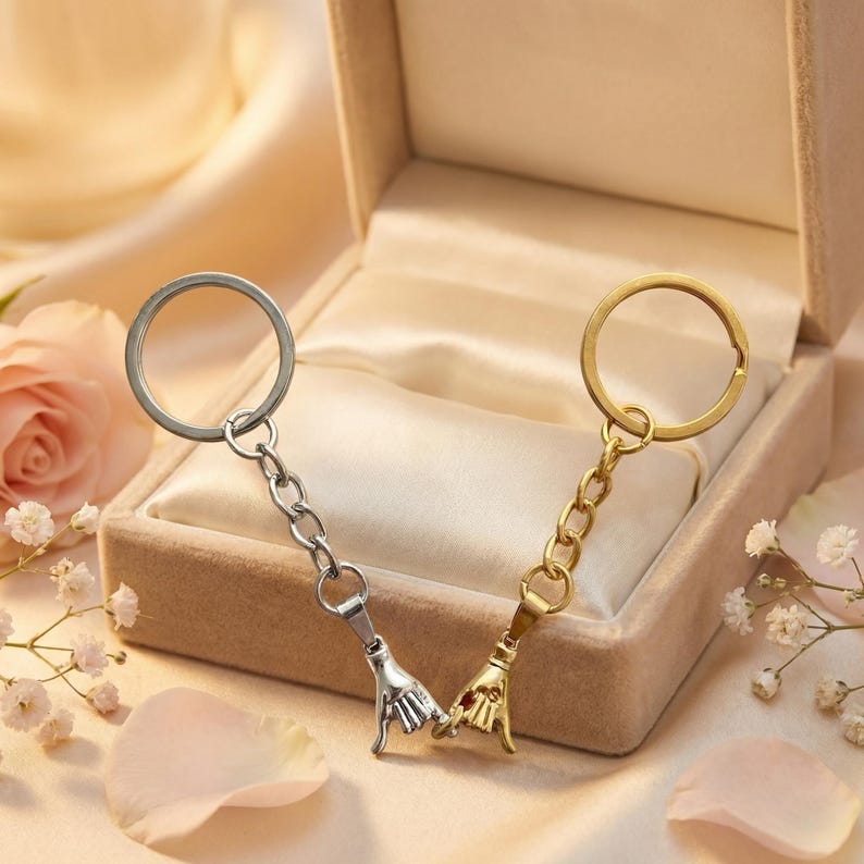 May include: Two keychains, one silver and one gold, each featuring a hand gesture charm. The charms are connected by a chain to a keyring. The keychains are presented in a small, open gift box on a satin surface.