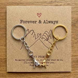 May include: A silver and gold keychain set with a promise hand gesture charm. The keychains are attached to a brown card with the words "Forever & Always" and a heart symbol. The card also includes text about a pinky promise.