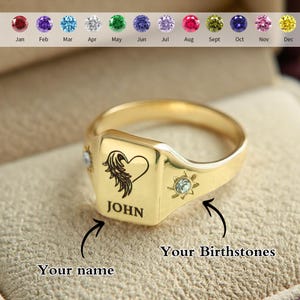 May include: A gold-colored signet ring with a square face, engraved with a heart and wing design, and the name "JOHN". The ring features a small gemstone on each side and a selection of birthstones.
