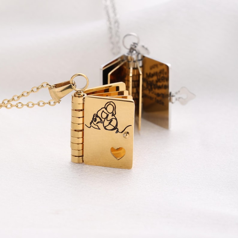 May include: A gold-colored book-shaped pendant necklace. The pendant is open, revealing pages with text. The front cover features a black line drawing of a mother and child, with a heart-shaped cutout. The necklace chain is silver.