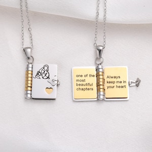 May include: Two silver necklaces with book-shaped pendants. One pendant is closed, with a family silhouette and heart cutout. The other pendant is open, with the words "one of the most beautiful chapters" and "Always keep me in your heart".