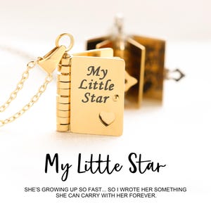 May include: A gold-coloured locket necklace featuring a book-shaped pendant. The pendant is inscribed with "My Little Star" and a heart. The necklace has a delicate chain. The text at the bottom says "My Little Star" and "SHE'S GROWING UP SO FAST... SO I WROTE HER SOMETHING SHE CAN CARRY WITH HER FOREVER."