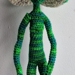 May include: A handmade crochet alien doll in shades of green and blue. The doll has large black eyes and a mushroom-shaped hat with a beige rim. The doll's arms and legs are slender, and the body is a solid green colour.
