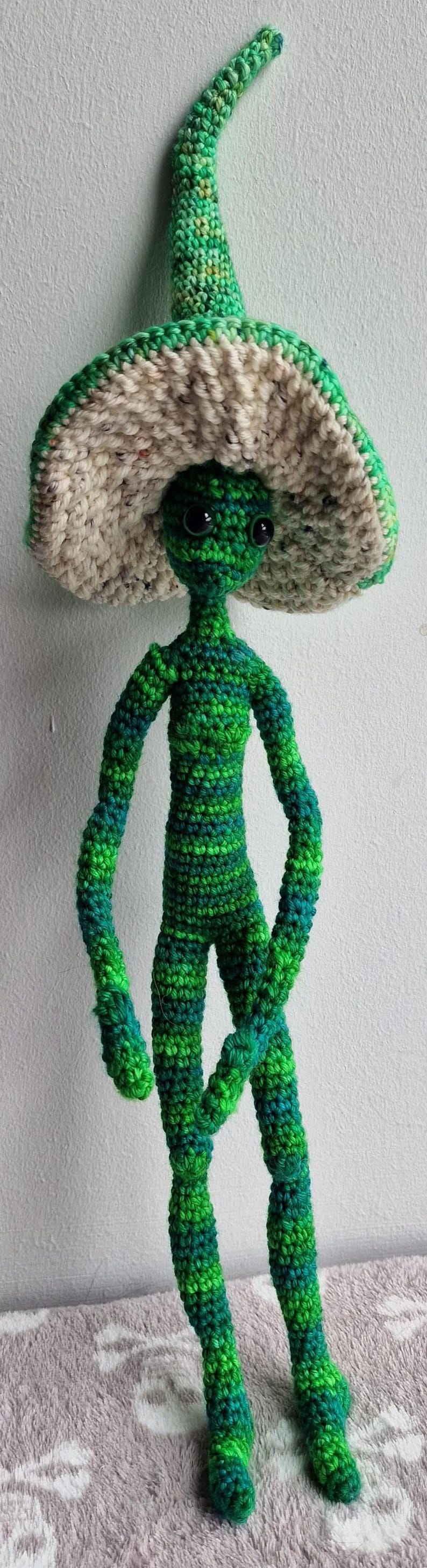 May include: A handmade crochet alien doll in shades of green and blue. The doll has large black eyes and a mushroom-shaped hat with a beige rim. The doll's arms and legs are slender, and the body is a solid green colour.