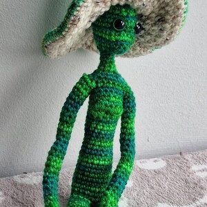 May include: A handmade crochet doll in shades of green and blue, with a large, floppy hat in green and cream. The doll has a slender body and is seated. The yarn has a variegated colour pattern.