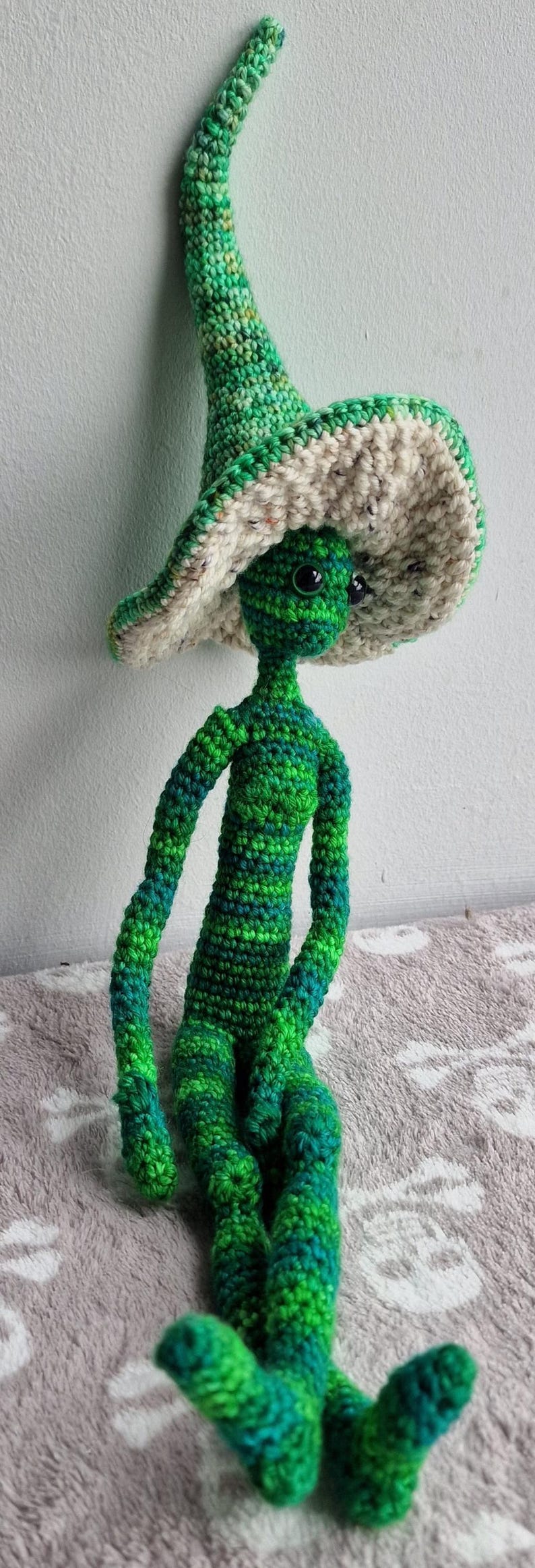 May include: A handmade crochet doll in shades of green and blue, with a large, floppy hat in green and cream. The doll has a slender body and is seated. The yarn has a variegated colour pattern.