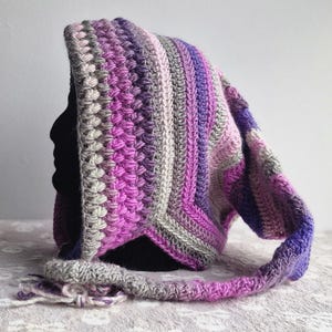 May include: A crocheted hooded cowl with a long, flowing scarf. The cowl is made in shades of purple, pink, and grey.