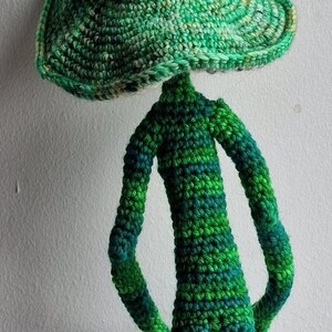 May include: A handmade crochet figure with a large, mushroom-shaped head and a slender body. The figure is primarily green with darker green and blue accents, and it stands approximately 30 cm tall. The whimsical design is perfect for home decor.