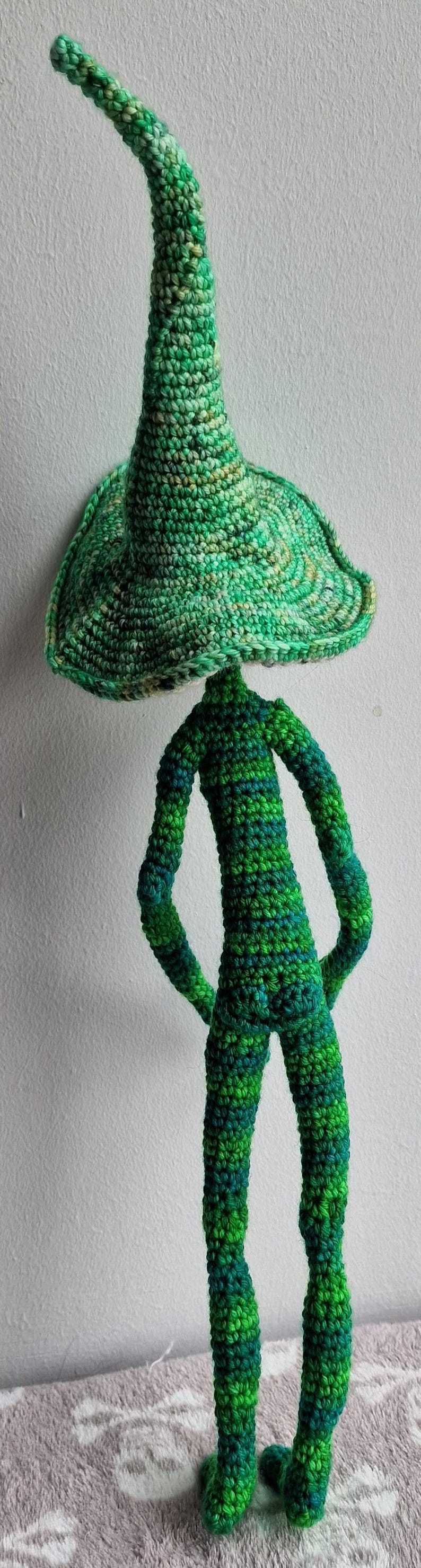 May include: A handmade crochet figure with a large, mushroom-shaped head and a slender body. The figure is primarily green with darker green and blue accents, and it stands approximately 30 cm tall. The whimsical design is perfect for home decor.