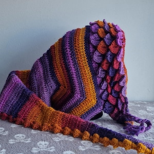 May include: A handmade crochet hood in shades of purple, orange, and red. The hood features a scale-like design on the top and a decorative edge. The scarf-like ends are long and finished with a textured border.
