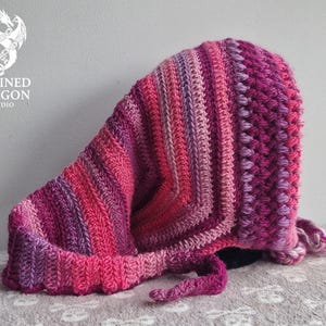 May include: A handmade, crocheted hood in shades of pink and purple. The hood features a textured stitch pattern and a long, pointed shape. The hood has a decorative edge and a tie closure. The Chained Dragon Studio logo is visible in the upper left corner.