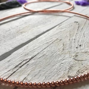 May include: Copper-colored wire jewelry, possibly a necklace or bracelet, with a twisted rope design. The jewelry is displayed on a weathered, white wooden surface. The image has a shallow depth of field, with other jewelry pieces in the background.