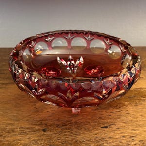 Könnte beinhalten: Eine rubinrote, geschliffene Glasschale mit einem gewellten Rand und drei kleinen Füßen. Die Schale weist ein detailliertes Muster aus Schliffmustern und kreisförmigen Aussparungen auf. Das Innere der Schale ist tiefrot, und die Schale steht auf einer Holzoberfläche.
