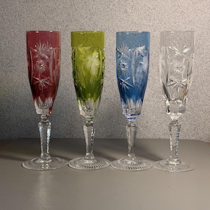 May include: A set of four tall champagne flutes with clear glass stems and bases. The bowls feature intricate cut glass star and floral patterns. The glasses are colored in deep red, vibrant lime green, bright blue, and clear transparent glass.