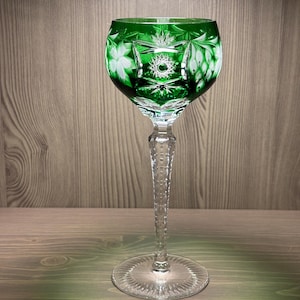 May include: A green cut-to-clear crystal wine glass featuring intricate starburst and floral patterns etched into the bowl. The stem is faceted with a clear glass finish, leading down to a circular base with a radial sunburst design at the center.