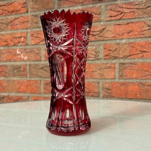 Könnte beinhalten: Eine rubinrote Kristallvase mit Sternen- und geometrischen Mustern. Die Vase hat einen ausgestellten Boden und einen gezackten Rand. Das geschliffene Glas reflektiert das Licht. Die Vase ist etwa 25 cm hoch.