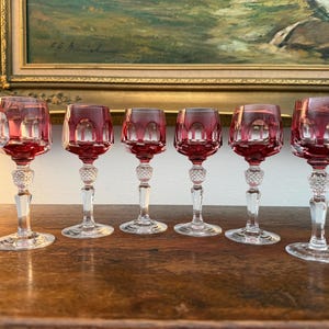 Op de afbeelding: Een set van zes kristallen glazen met dieprode kelken met gefacetteerde slijppatronen en heldere stelen. Elk glas heeft een textuurrijk, diamantgeslepen knopdetail boven een heldere ronde voet, consistent over alle zes de glazen.
