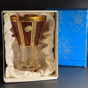 Può includere: Un vaso in vetro di Boemia color rubino con finiture dorate, presentato in una scatola bianca con carta velina. Il vaso ha un design inciso dettagliato e un bordo dorato. È visibile una scatola blu con dettagli dorati.